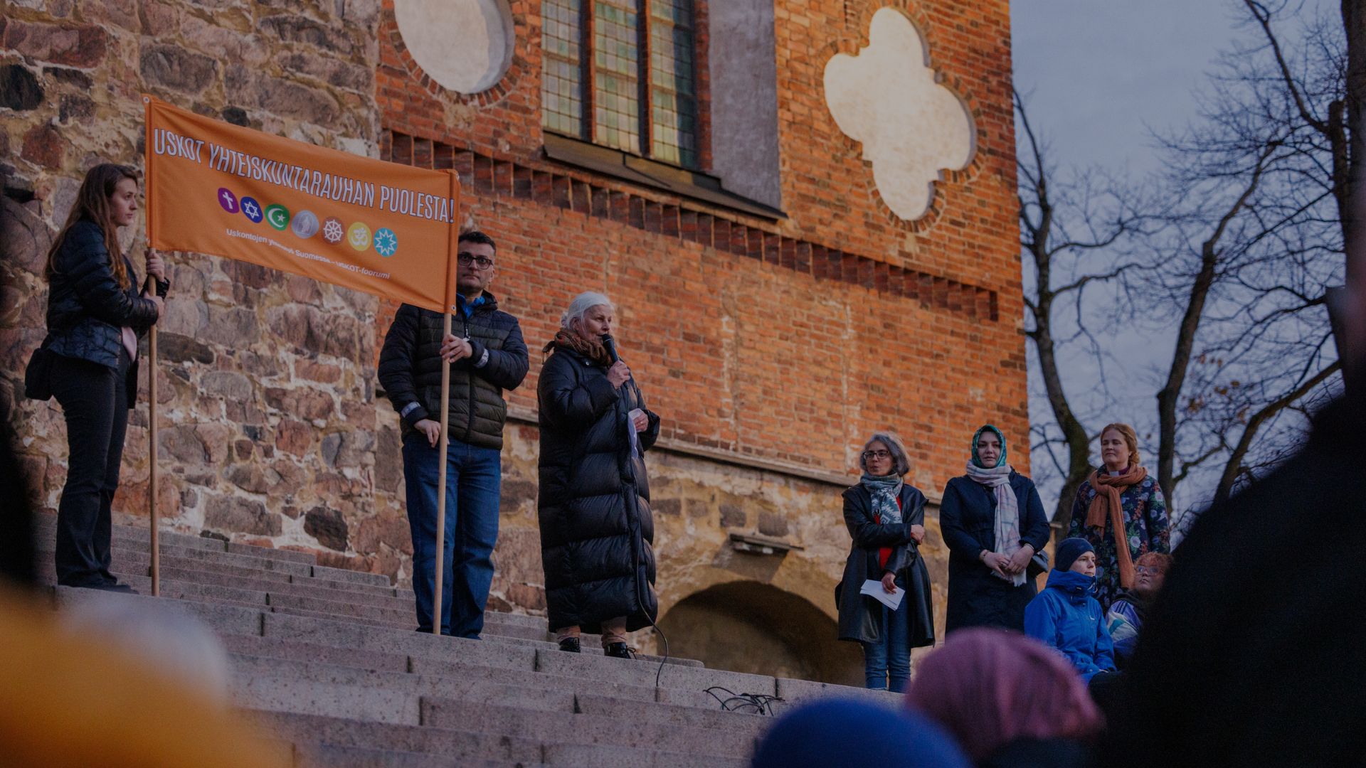 Kuvituskuvassa uskontojen rauhankävelyltä, jossa henkilö puhuu mikrofoniin ulkona Turun tuomiokirkon portailla kahden henkilön pitäessä vieressä kylttiä "uskot yhteiskuntarauhan puolesta"